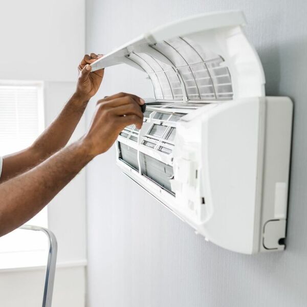 An individual in a blue hard hat looking into a airconditioning unit mounted on the wall